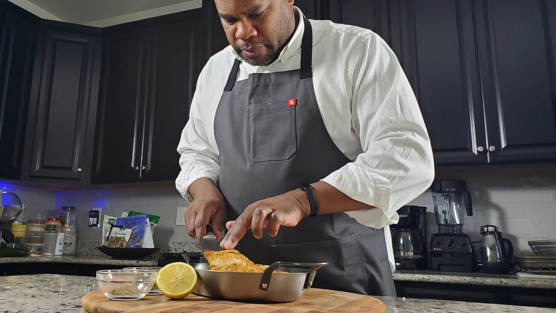 A man in a gray apron slices cooked fish in a pan on a wooden cutting board in a modern kitchen, with a lemon and glass bowls nearby.