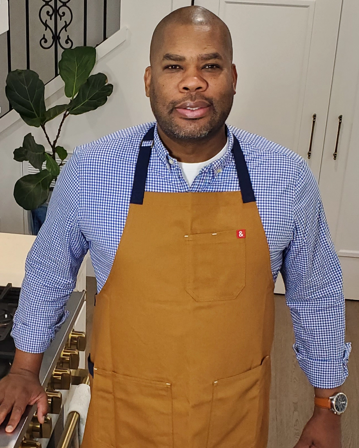 A confident man in a blue checked shirt and mustard apron stands in a bright home kitchen, with white cabinets and a leafy plant behind him.
