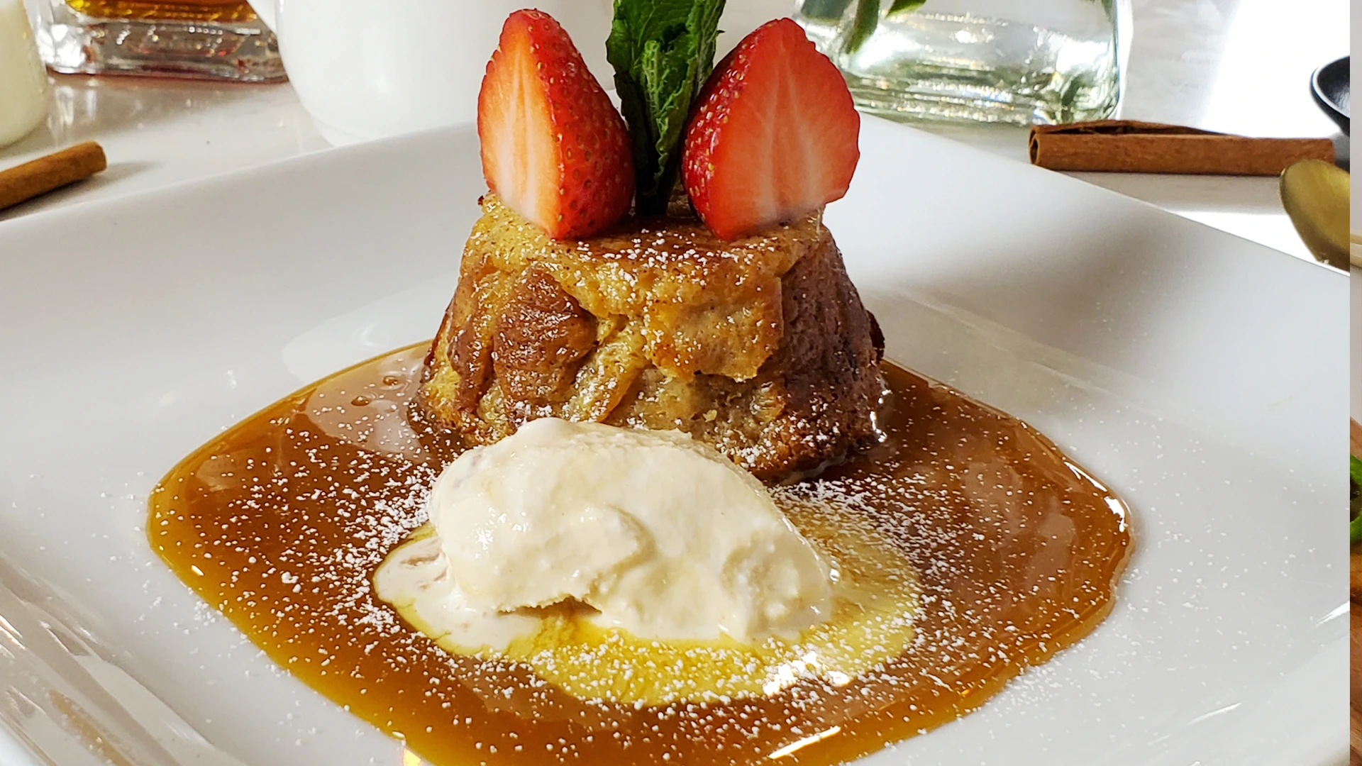 A warm bread pudding sits in a pool of caramel sauce with vanilla ice cream, topped with strawberry halves and mint on a white plate; inviting and indulgent.