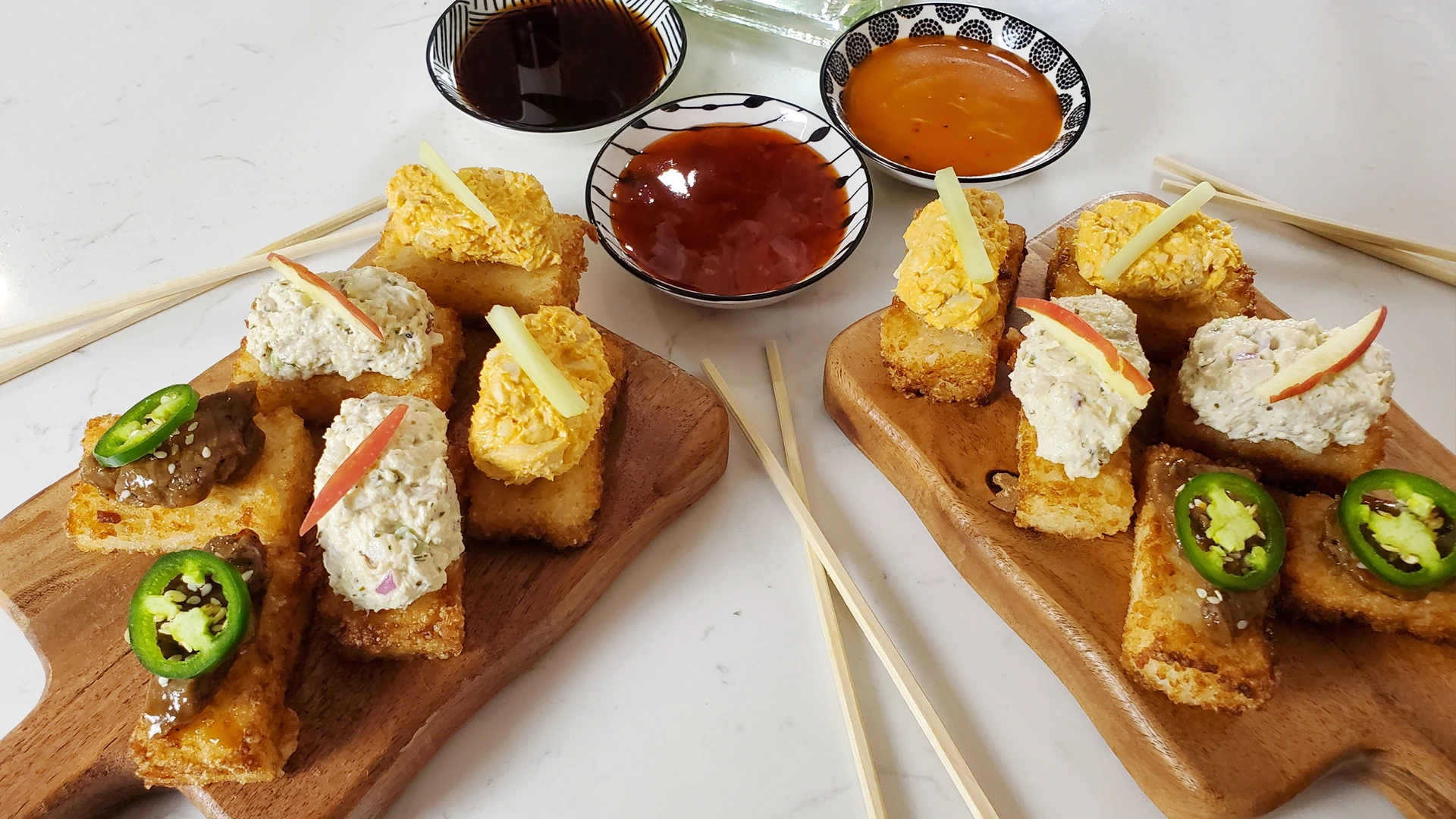 Two wooden platters hold assorted bite-sized fried appetizers topped with jalapeño, creamy fillings, and cheese, with three dipping sauces behind them. The spread looks inviting.