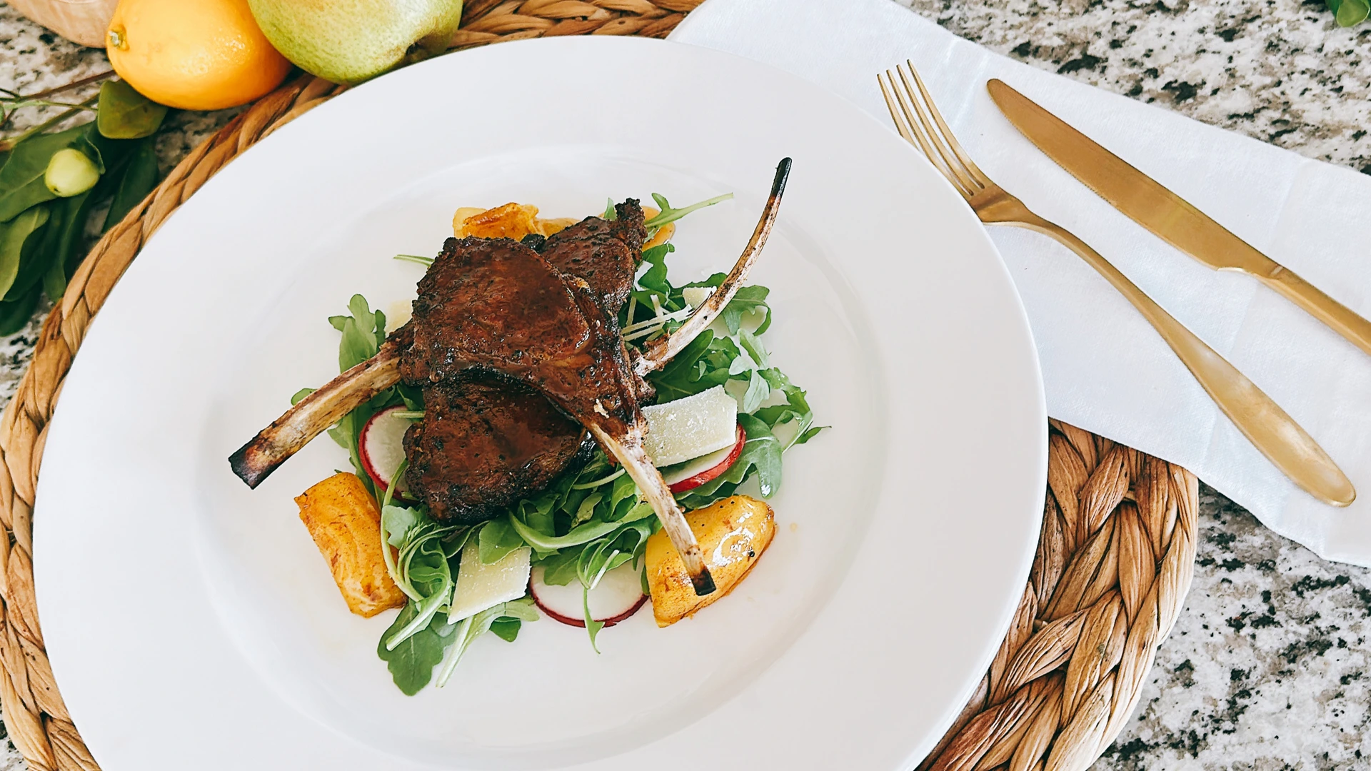 Three seared lamb chops rest on a fresh green salad with fruit wedges on a white plate, beside gold cutlery and a fruit basket; an inviting meal.
