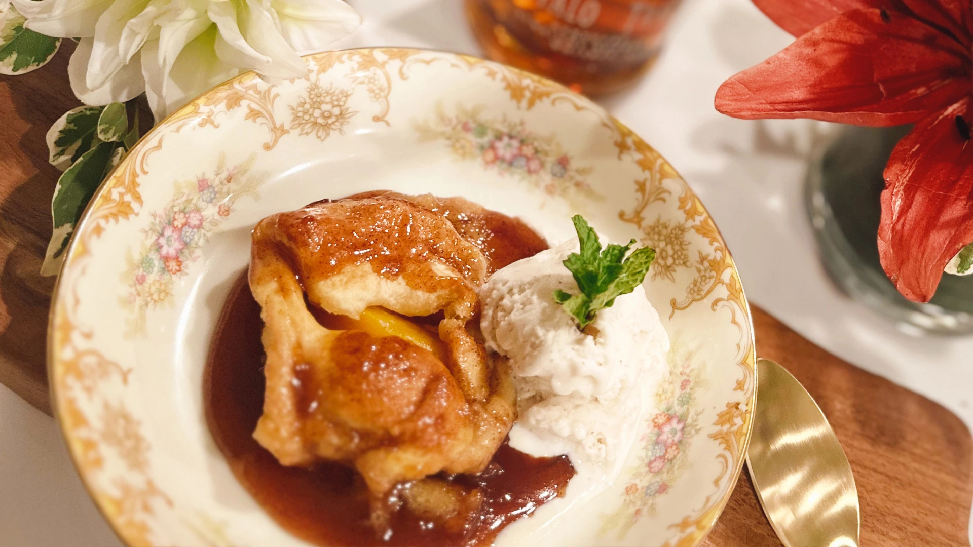 Golden-brown peach dumplings with caramel sauce and whipped cream on a floral china plate, framed by white flowers and a red lily for a warm, elegant dessert setting.