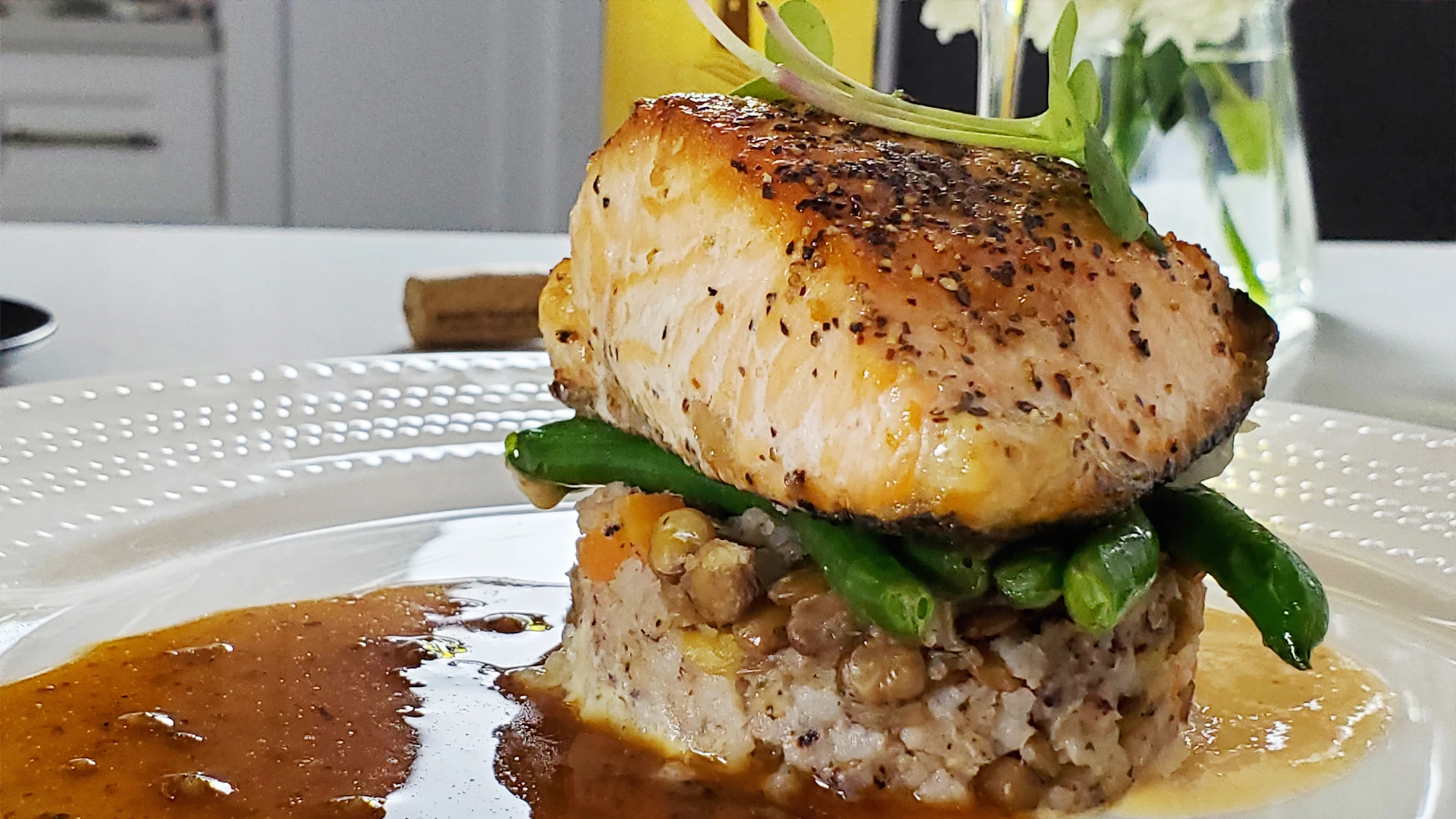 A golden, seared salmon fillet rests on lentils and grains with fresh greens, surrounded by glossy brown sauce on a white plate; appetizing, elegant presentation.
