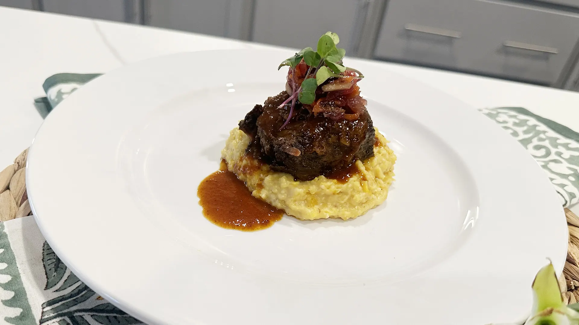 A braised beef short rib sits on creamy yellow grits, topped with fresh microgreens and glossy brown sauce on a large white plate.
