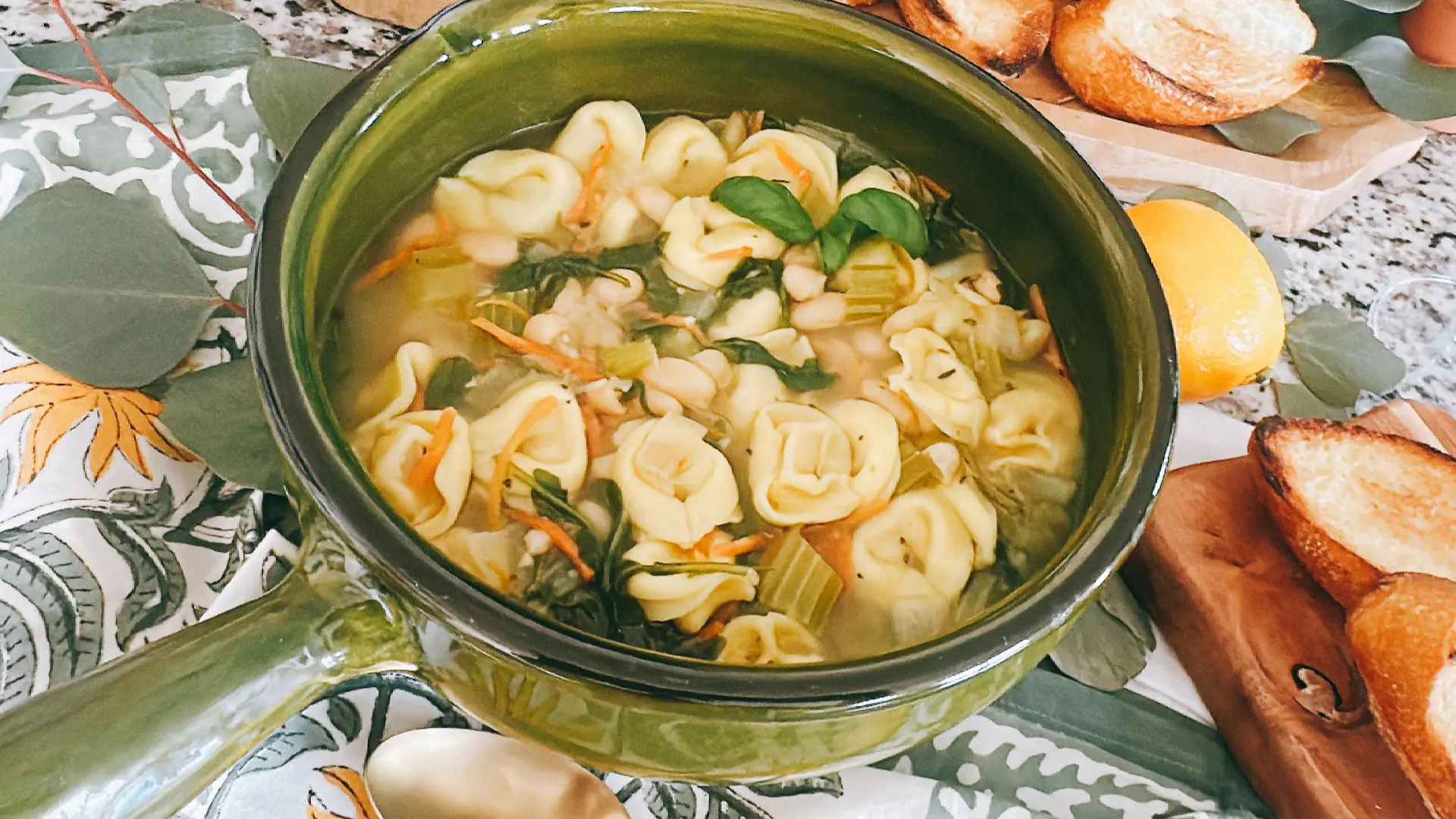 Overhead view of a green pot filled with tortellini soup, vegetables, and herbs, surrounded by sliced bread on a patterned tablecloth; warm, homey meal.